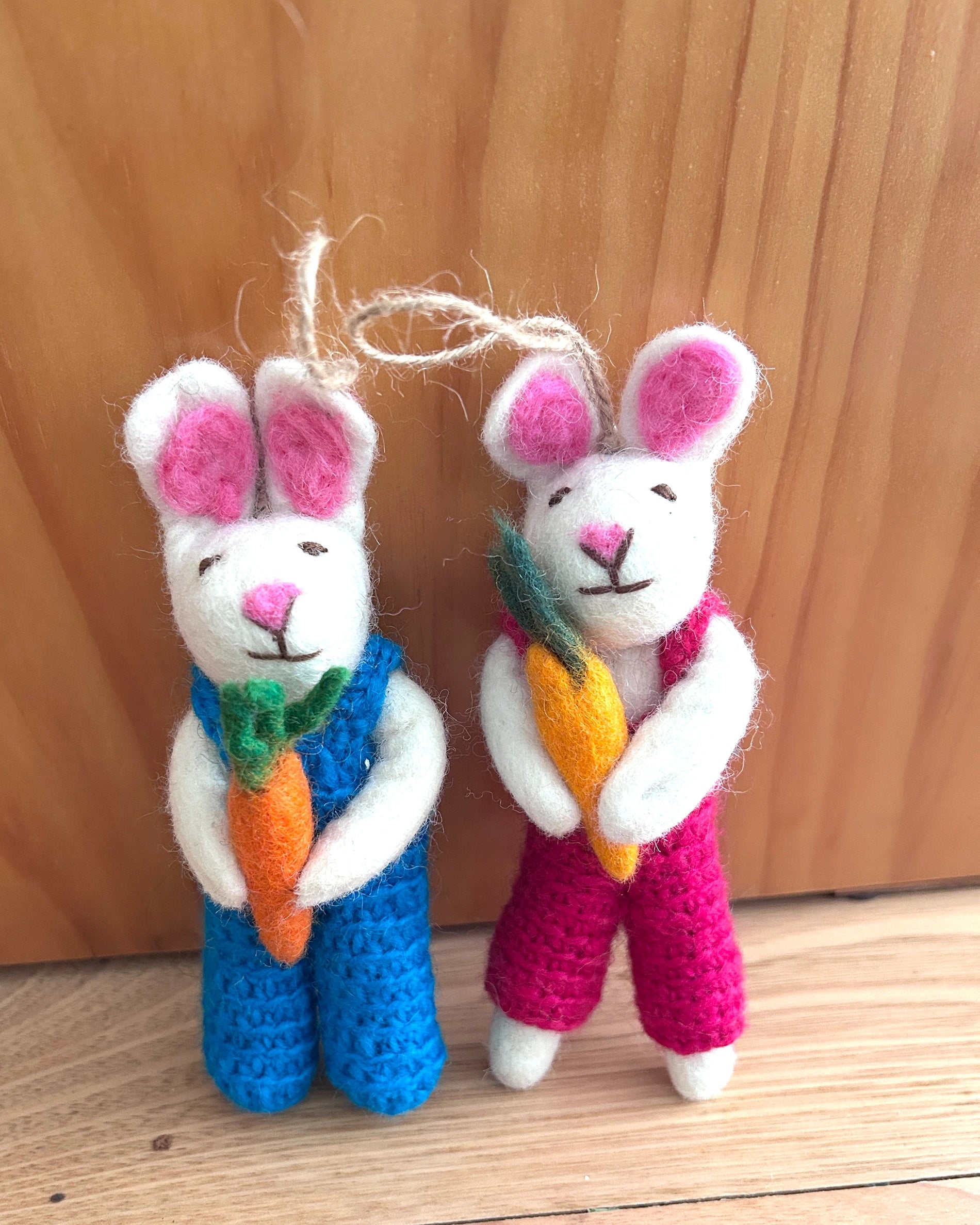Two knitted or crocheted bunnies, one in blue and one in pink, holding carrots against a wooden background.