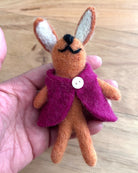 Small felt bunny toy wearing a purple coat held in a hand on a wooden surface