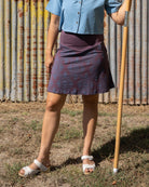 Woman in a blue shirt and patterned skirt holding a yellow umbrella against a corrugated metal wall.