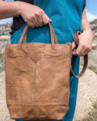 Person holding a brown leather tote bag in a rocky outdoor setting