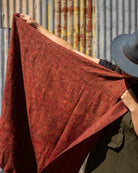 A person wearing a dark hat holds up a large rust-red woven , shawl, or scarf, against a corrugated metal wall with rust patches.