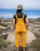 Person wearing yellow overalls and a striped hat standing on rocky terrain with a scenic background.