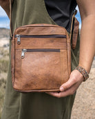 Brown leather crossbody bag held by a person in an outdoor setting