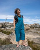 Person wearing a teal overall standing on a rocky outcrop with a blue sky and mountains in the background