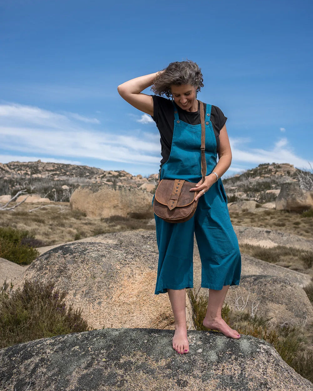Woman in a blue dress overalls with a leather bag on rocky terrain with a blue sky.