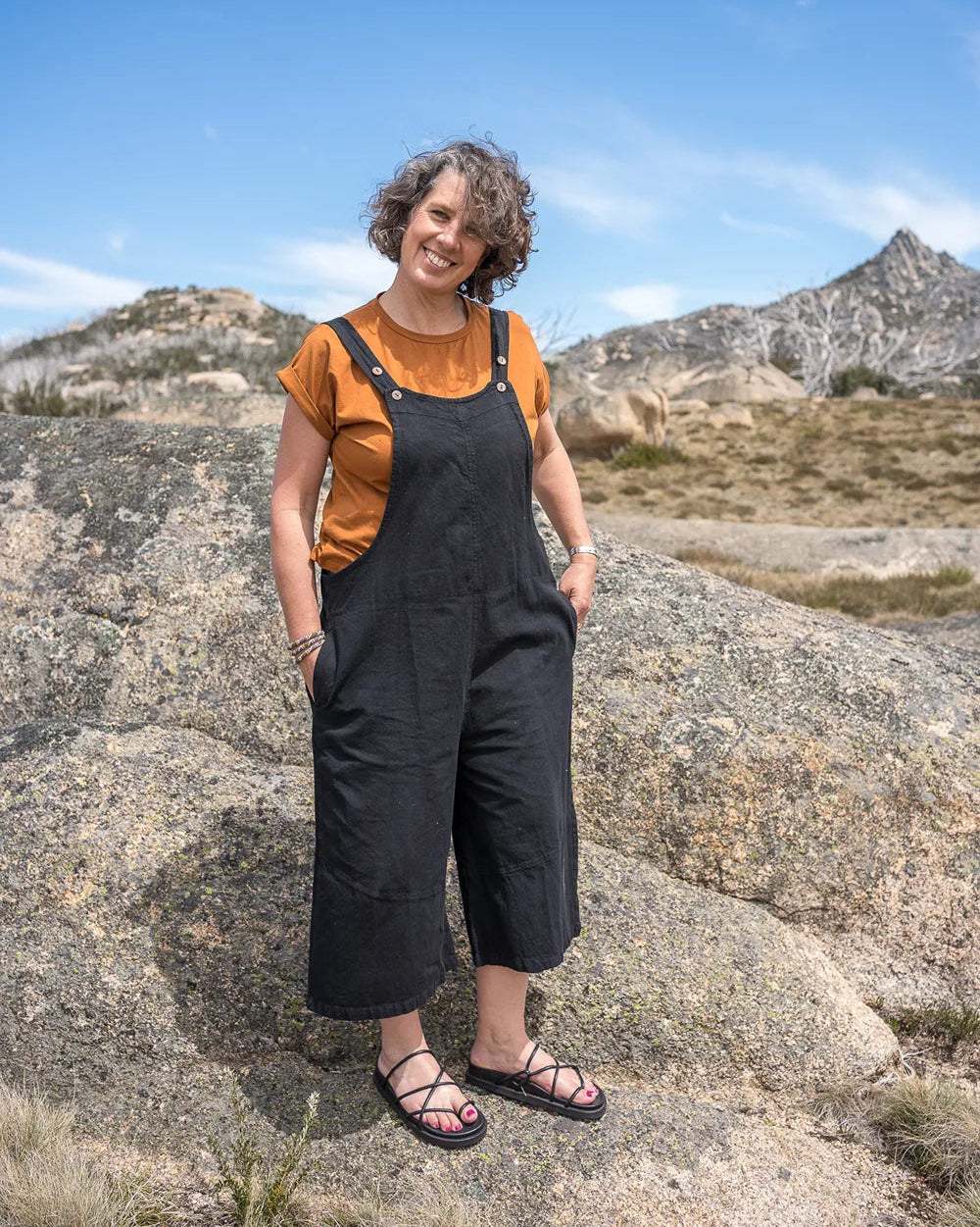 Woman in black overalls and orange shirt standing on rocky terrain with mountains in the background