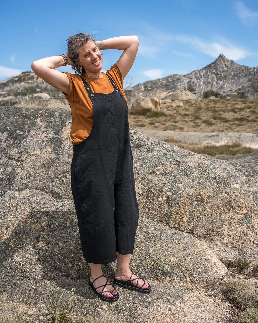 Woman in black overalls and orange shirt standing on a rocky landscape with mountains in the background