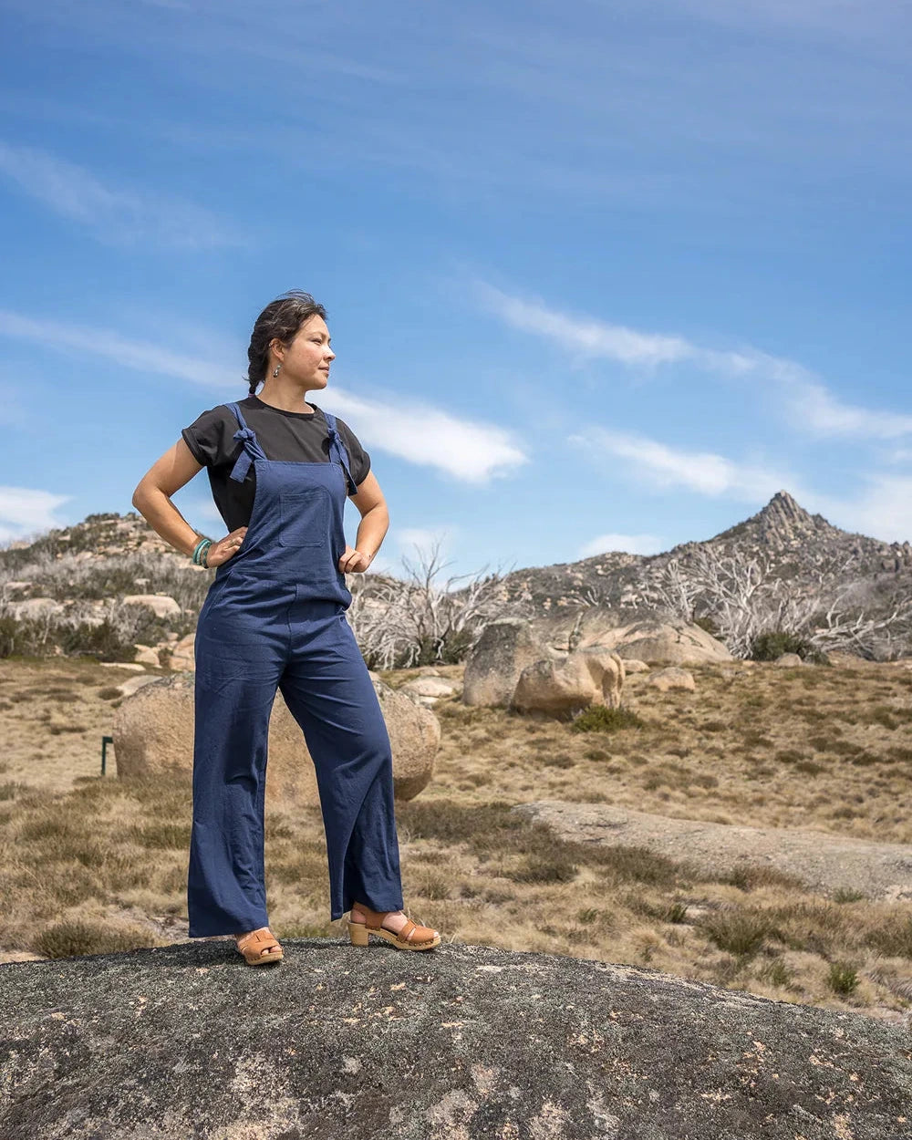 Person wearing a blue jumpsuit standing on a rocky outcrop with mountains in the background