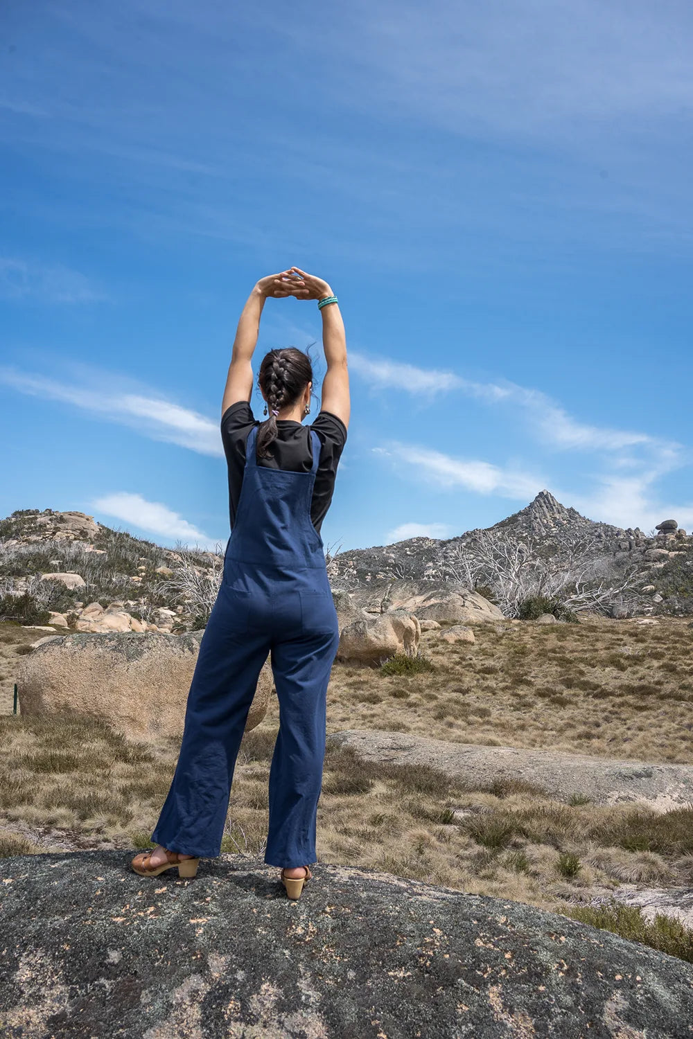 Person in blue overalls standing on a rock with mountains and clear sky in the background