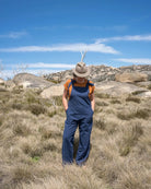 Person wearing a hat and blue overalls standing in a rocky, grassy landscape with a clear blue sky.