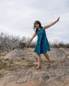 Woman in a blue dress standing on rocks with arms outstretched in an open landscape