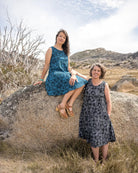 Two women in patterned dresses sitting on a rock in a natural landscape.