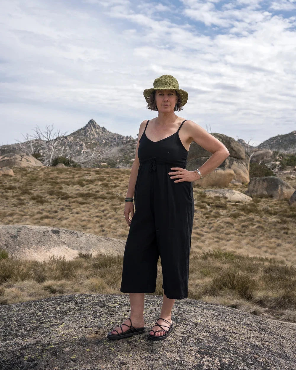 Woman in a black jumpsuit and hat standing on a rocky outcrop with mountains in the background