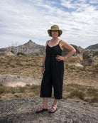 Woman in a black jumpsuit and hat standing on a rocky outcrop with mountains in the background