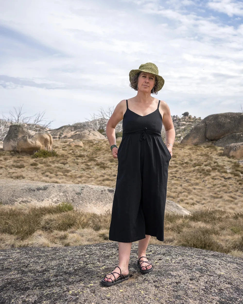Woman in a black jumpsuit and hat standing on a rocky landscape with a blue sky.