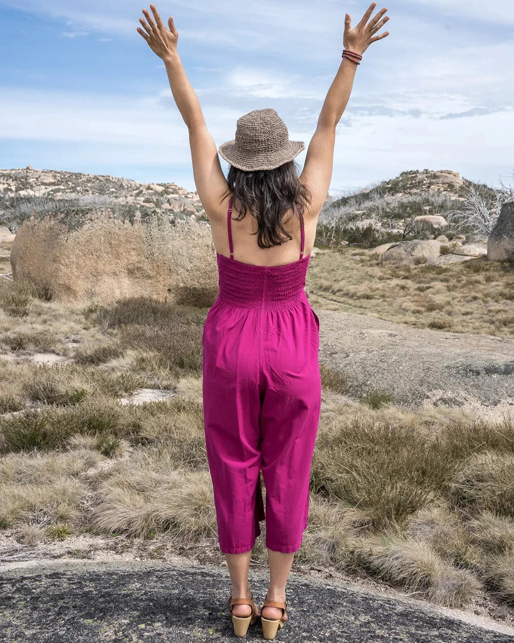 Woman in a pink jumpsuit and hat standing with arms raised in a natural landscape.