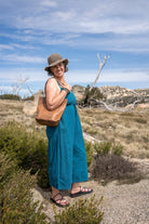 Woman in a blue dress and hat standing in a desert landscape with a brown bag.