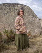Person wearing a patterned jacket and green skirt standing in front of a large rock with a blue sky.