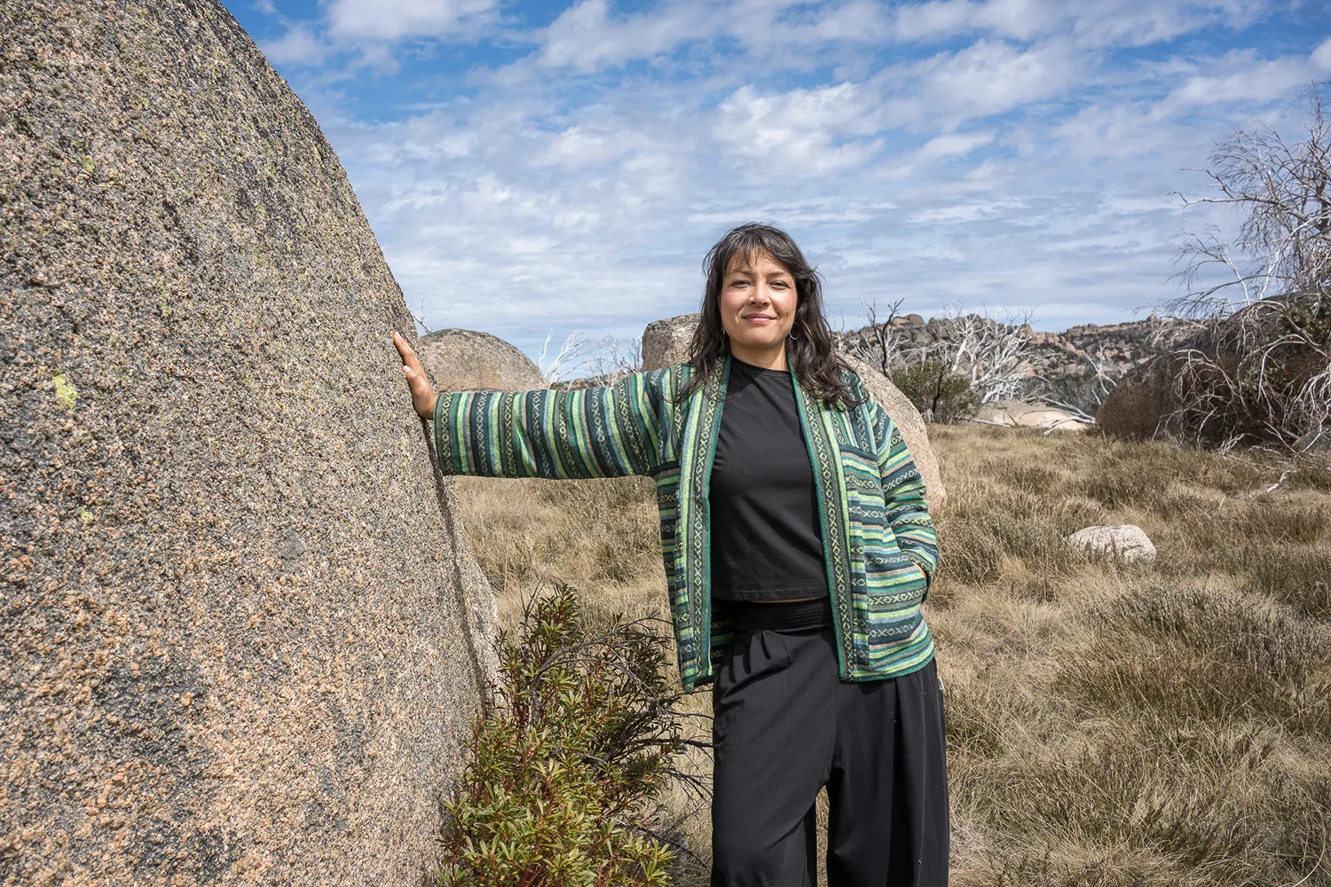 Person standing next to a large rock in a natural landscape with blue sky and clouds.