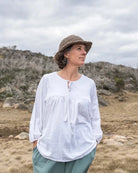 Woman in a white blouse and green pants standing on a rocky outcrop with a mountainous landscape in the background.