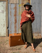 A woman wearing a wide-brimmed black hat, a rust-red shawl, and an olive green midi dress stands outdoors.