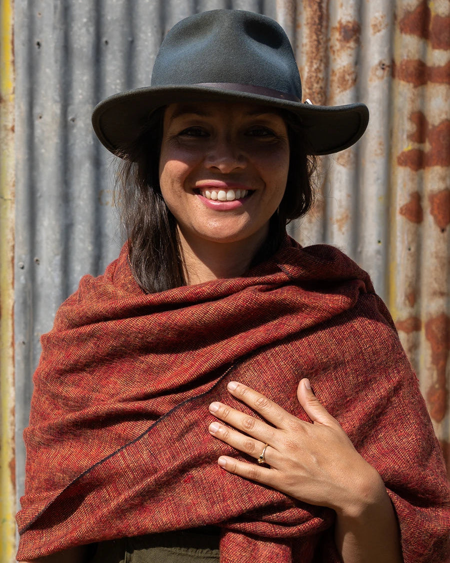 A woman wearing a wide-brimmed dark hat and a rust-red textured shawl or wrap, standing in front of a corrugated metal wall.