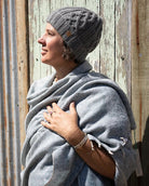 A woman wearing a gray cable-knit beanie and a light gray shawl wrapped around her shoulders, standing against a rustic corrugated metal and wooden wall.