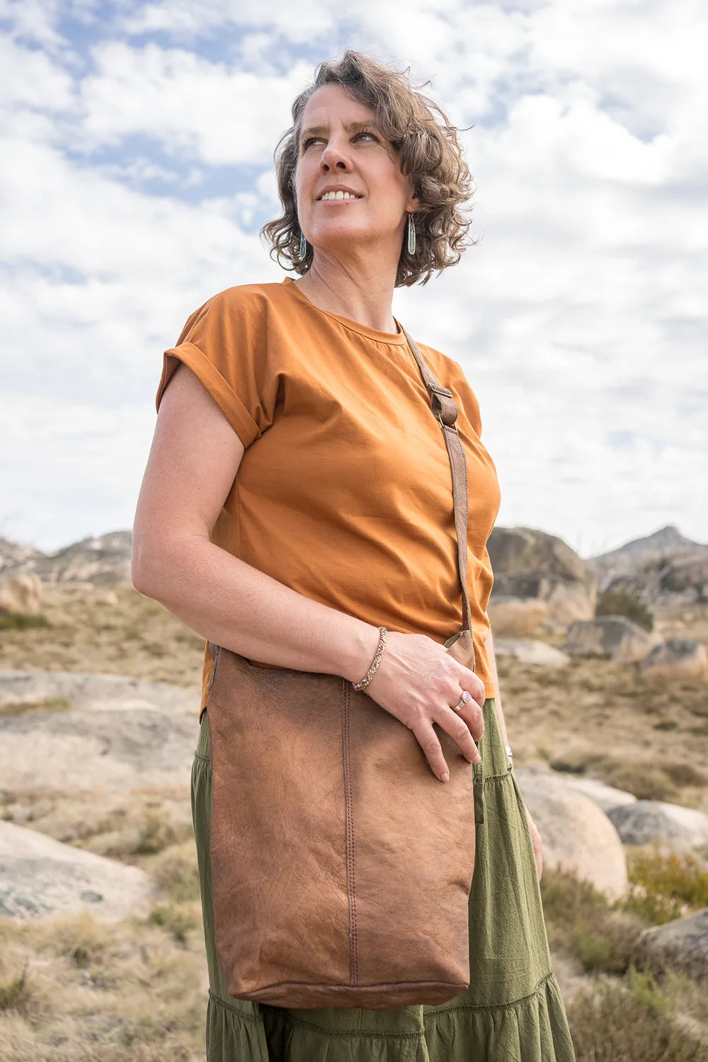 Woman in an orange shirt and brown apron standing in a rocky landscape with mountains in the background.