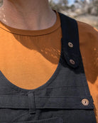 A close-up of a person outdoors wearing dark charcoal gray canvas bib overalls over a mustard orange t-shirt. The overalls feature a bib front with buttoned strap details and a chest pocket, photographed against a blurred rocky desert background.