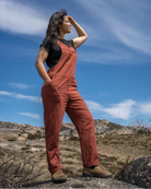A woman stands outdoors in a rocky, grassy landscape under a blue sky, wearing rust-orange bib overalls over a black short-sleeve top and brown boots.