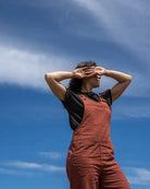 A woman wearing rust-brown overall over a black t-shirt stands against a bright blue sky with wispy white clouds, shielding her eyes with her hands.