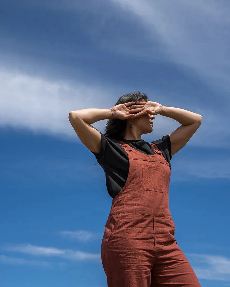 A woman wearing rust-brown overall over a black t-shirt stands against a bright blue sky with wispy white clouds, shielding her eyes with her hands.