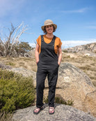 A woman wearing black utility overalls over an orange t-shirt, a straw hat, and sandals, standing on a large rock in an outdoor rocky landscape under a blue sky.