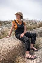 A woman wearing a straw hat, a mustard yellow short-sleeve top, and black overall-style pants sits on a large rock in an outdoor rocky landscape under a bright sky.