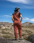 A woman stands on a rocky hillside in a dry grassy landscape, viewed from behind, wearing rust-colored overalls over a black t-shirt, a dark wide-brim hat, and boots, with a blue sky in the background.