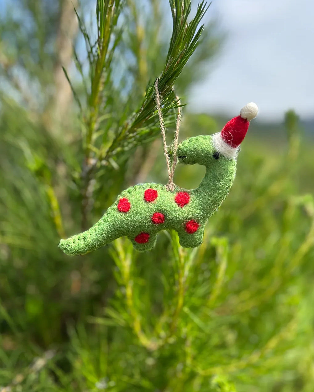 Green felt dinosaur ornament with red spots and a Santa hat hanging on a tree.